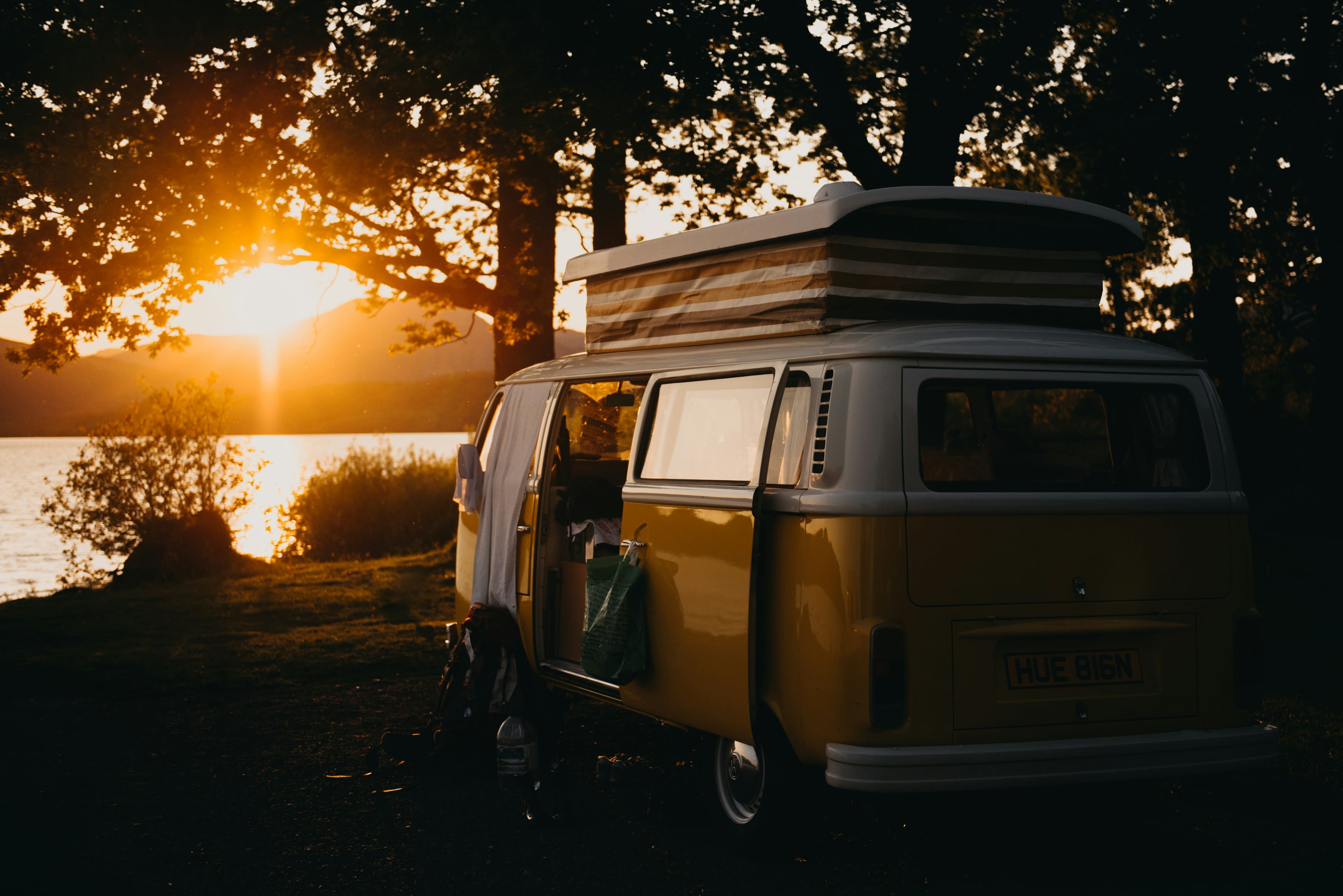 A yellow and white VW camper van parked by a loch at sunset, its pop-top raised.